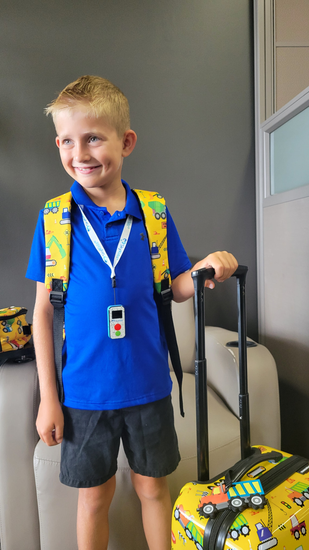 Smiling young boy wearing a Sky Kidz GPS phone device on a lanyard with his backpack and suitcase