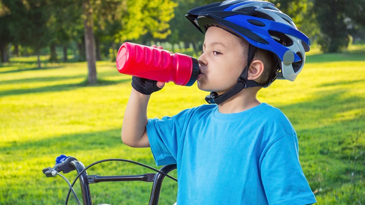 Young boy in a bike helmet drinking water beside his bicycle in a sunlit park